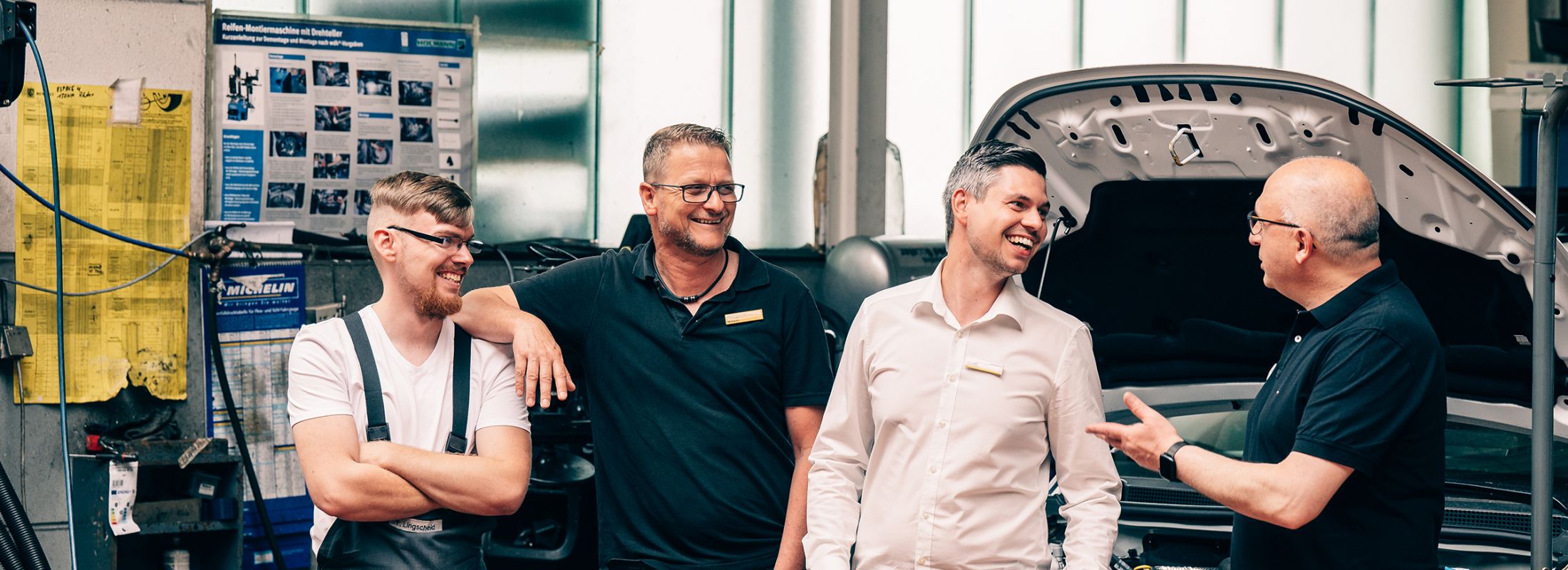 Vier Männer stehen in einer Werkstatt neben einem offenen Motorraum und unterhalten sich