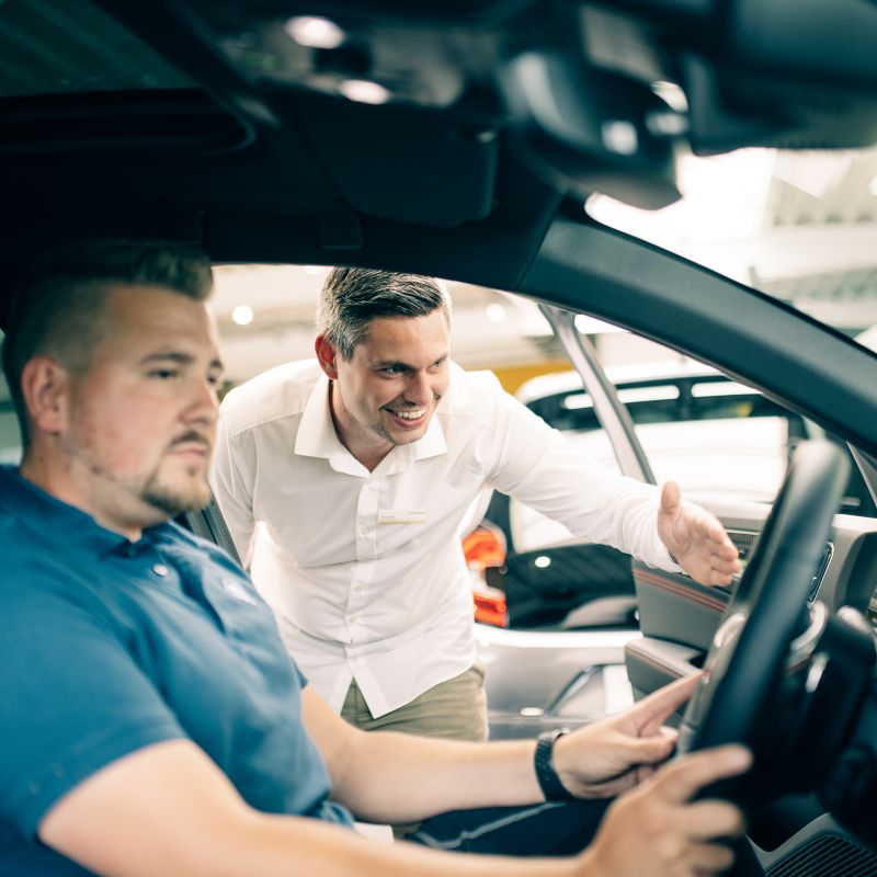 Ein Mann sitzt im Auto während ein Verkäufer ihm etwas im Innenraum zeigt