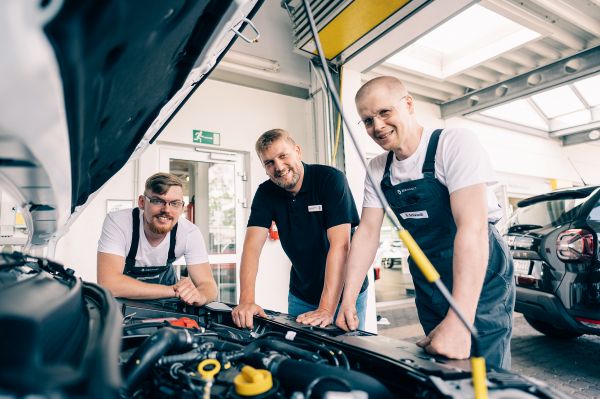Drei Mitarbeiter stehen in einer Werkstatt und schauen gemeinsam in den Motorraum eines Autos
