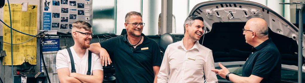 Vier Männer stehen in einer Werkstatt neben einem offenen Motorraum und unterhalten sich