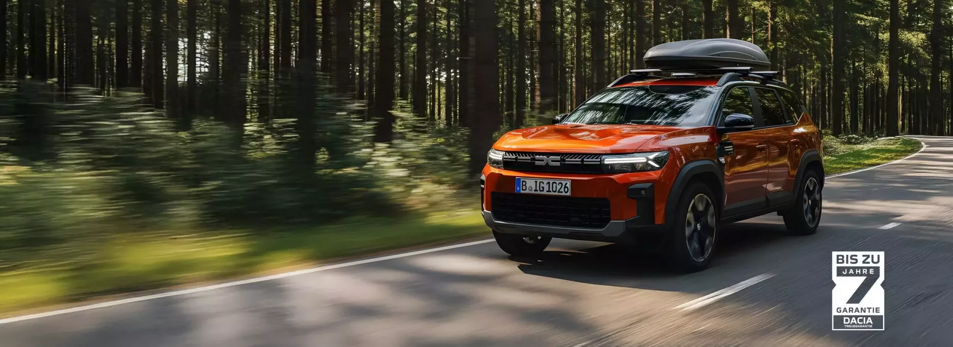 Oranger Dacia SUV mit Dachbox fährt auf einer Straße durch einen Wald