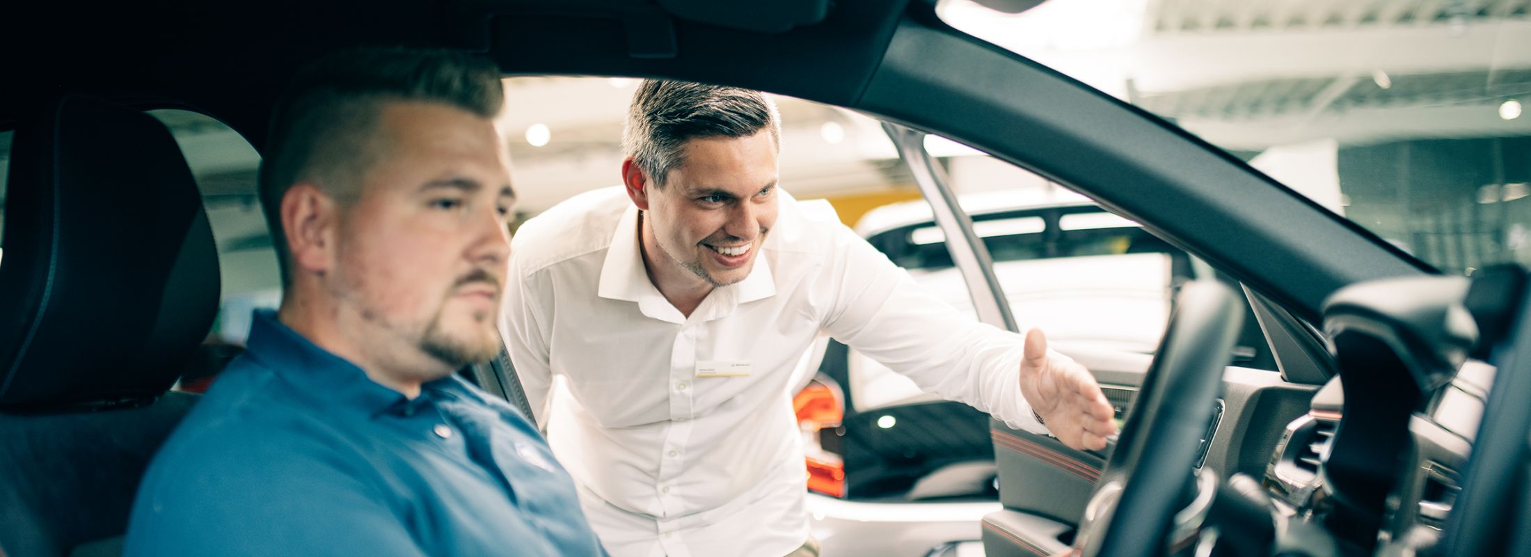 Ein Mann sitzt im Auto während ein Verkäufer ihm etwas im Innenraum zeigt